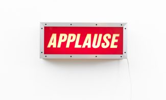 A sculpture of a wall aluminum lightbox with the word "APPLAUSE" in white slanted lettering on a red background and a white cord hanging down from the right side, mounted on a white wall.