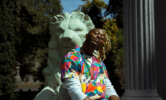 A photo portrait of Cliff Notez, an African-American musician in a colorful shirt leaning against a lion statue with a roman column and greenery in the background.