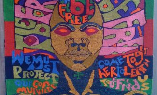 A colorful textile depicting a face with brown skin and red eyes surrounded by words including "To be free."