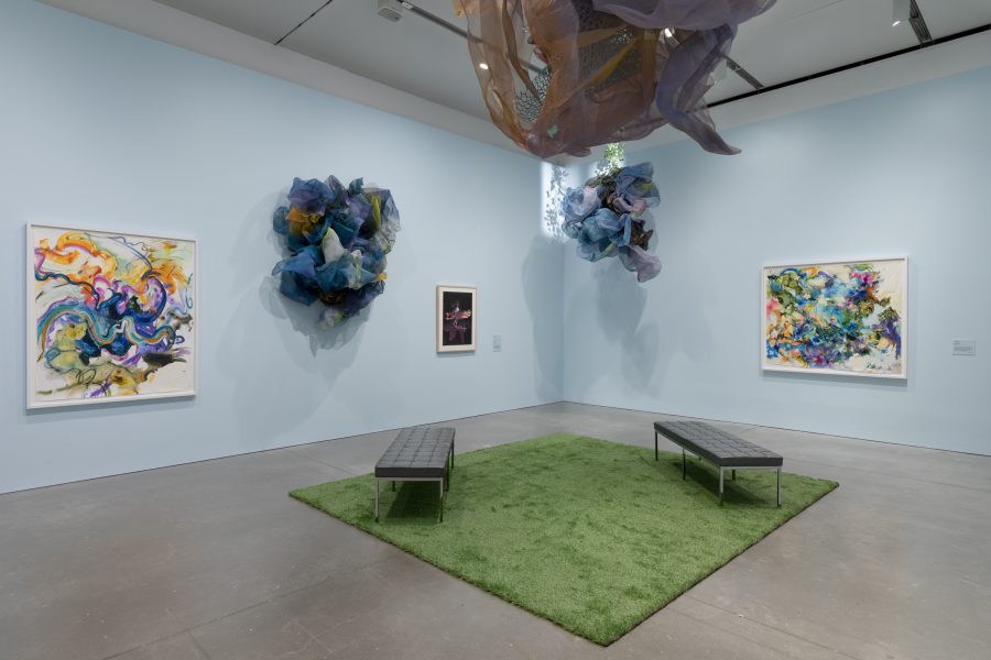 Blue gallery with green rug and paintings on wall and suspended fabric sculptures