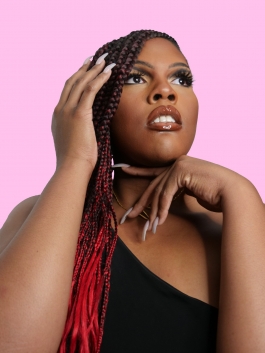 A glamour head shot of an African-American femme person with red and black braids, posing with their hands in front of a pink backdrop.