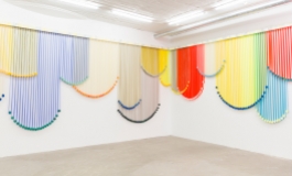 Along two adjacent white walls, a series of colorful hanging elements form geometrically shaped curtains.
