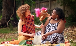 Two adult women interact with each other outdoors, smiling, with a stuffed chicken