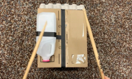 An aerial image of a pair of hands holding sticks while striking a DIY drum made of a box, a milk carton, and an egg carton.