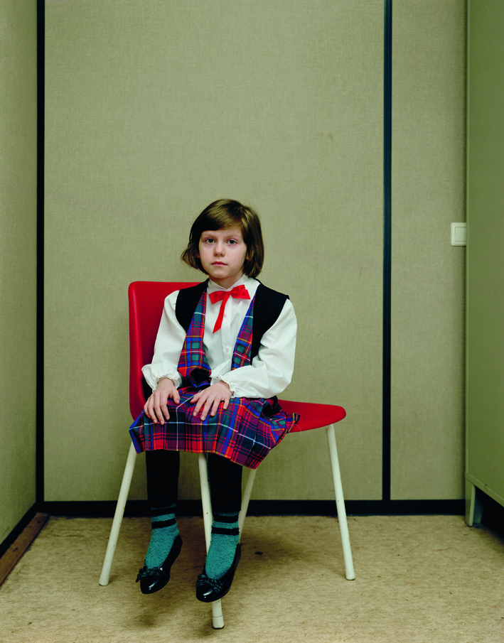 A color photograph of a young light-skinned girl with short brown hair in a blouse, vest, plaid skirt, and dress shoes sitting on a red chair with her feet not touching the ground, positioned at an angle and gazing at the viewer.
