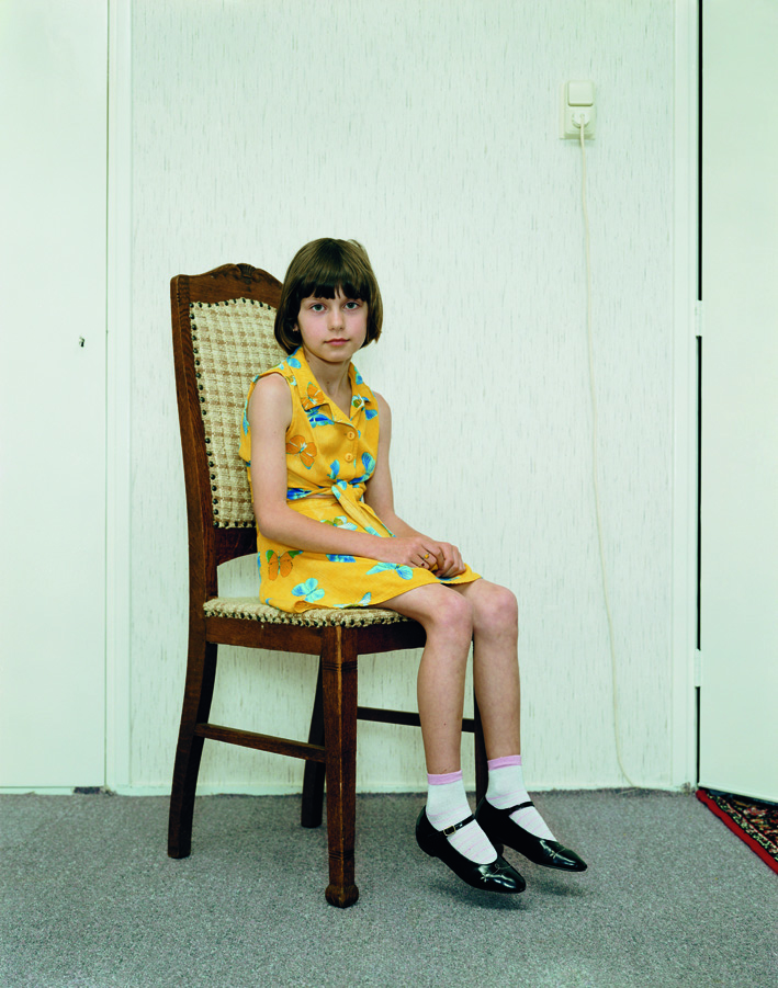 A color photograph of a light-skinned girl with short brown hair in a yellow dress, mary janes, and white socks sitting on a wooden chair, positioned at an angle and gazing at the viewer.