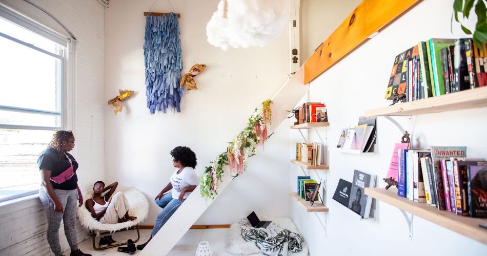 Three dark skinned femmes talk in relaxed manner in a brightly lit room  with shelves of books, plants, and hanging installations. 