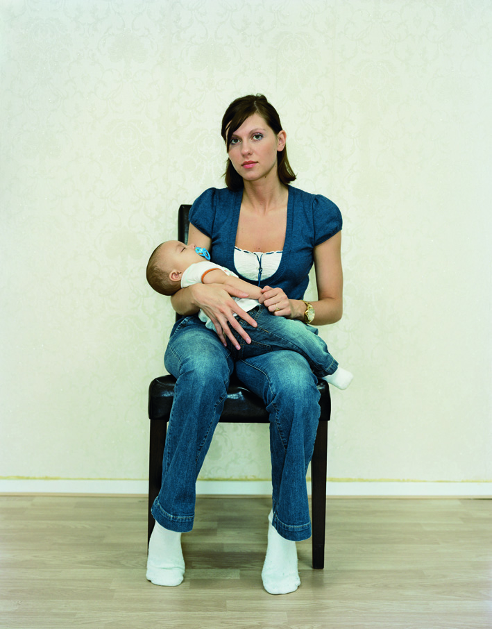 A color photograph of a light-skinned woman with short brown hair in a dark top, jeans, and white socks sitting in a chair with a baby in her arms, facing and gazing at the viewer. 