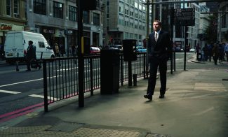 A color photograph of a light-skinned man in a suit walking along an urban street.