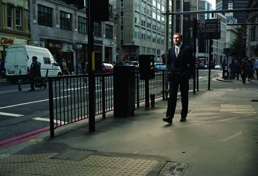A color photograph of a light-skinned man in a suit walking along an urban street. 