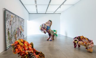 A multi-colored, glittery horse sculpture is in the middle of the room. A few smaller, similarly textured sculptures are partially in view, and a collaged tapestry hangs to the left while a multi-colored circular collage hangs in the very back.