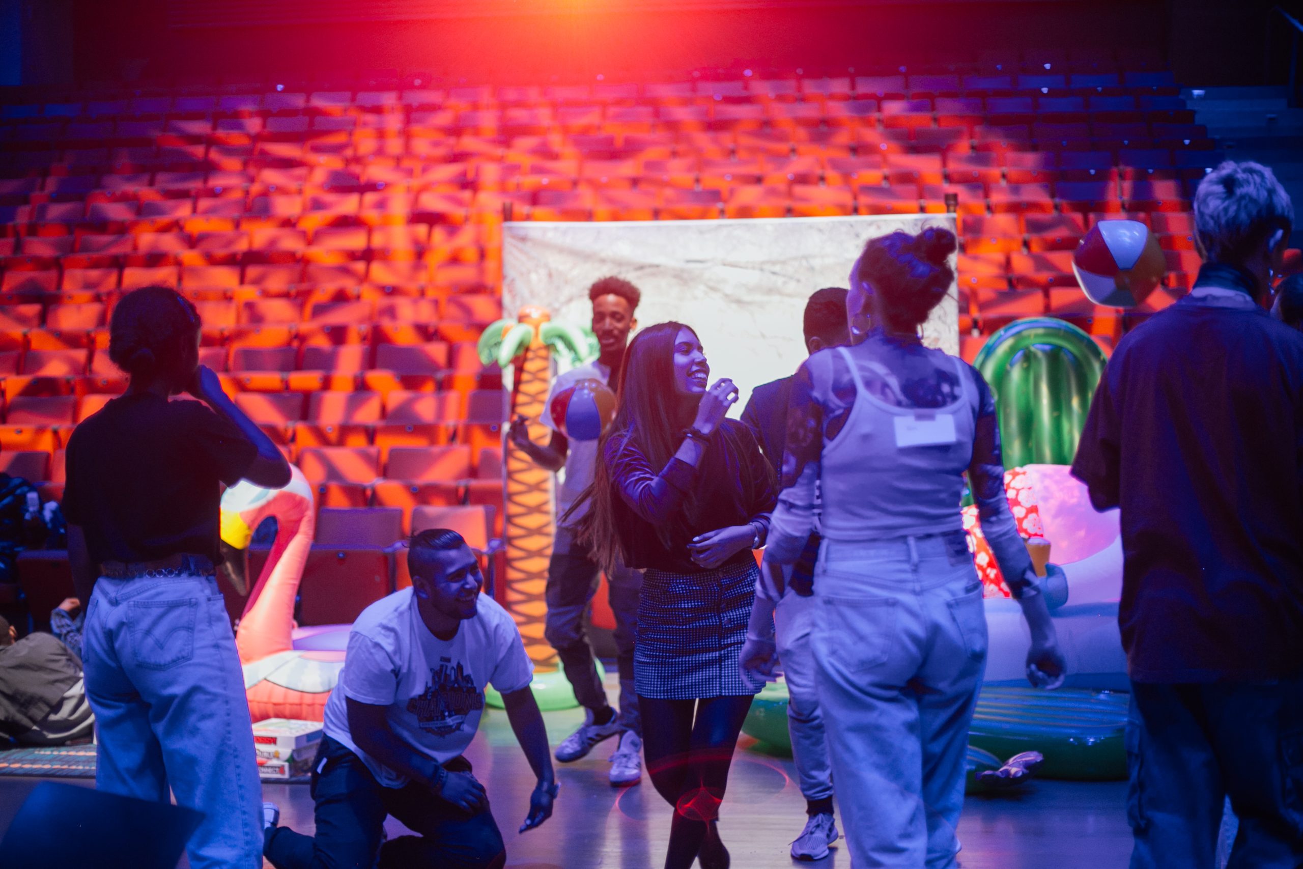 A group of teenagers socialize at the floor of a theatre, standing in front of a photoshoot display with tropical-themed props.