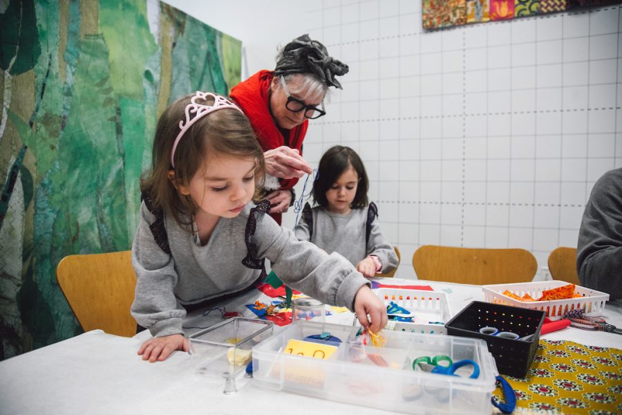 Two young girls in matching sweatshirts make art with artist Merill Comeau, who wears large glasses, a red scarf, and a cloth tied around her gray hair.