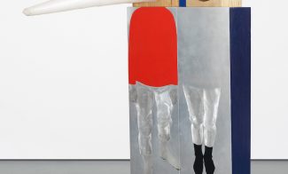 A sculpture comprised of a rectangular solid painted to appear as two figures standing side by side. Their faces appear on wooden boxes atop the structure: one as a long white triangular cone, one a naturalistic face painted on a flat circle.