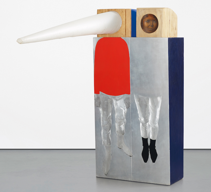 A sculpture comprised of a rectangular solid painted to appear as two figures standing side by side. Their faces appear on wooden boxes atop the structure: one as a long white triangular cone, one a naturalistic face painted on a flat circle.