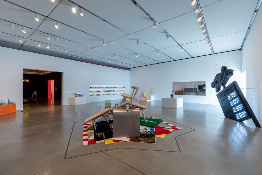 A gallery with a heap of rugs and furniture in the middle, a sculpture resembling a tilted all-black gas station sign on the right, and three shelves of sneakers in the background.