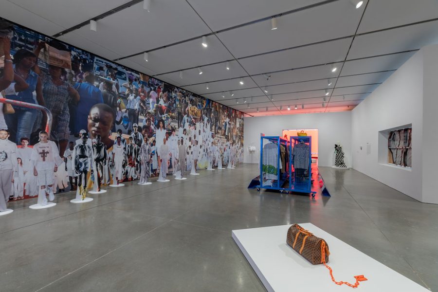 A gallery with a wall-sized photo collage of an African street scene and dozens of models in all white, with standing model cutouts in front, racks of clothing, and a Louis Vuitton bag bound to a plinth by a neon orange chain.