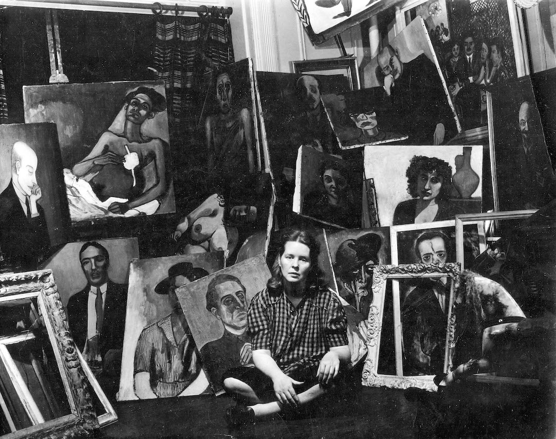 A black and white image of painter Alice Neel sitting down and surrounded by her painting portraits.