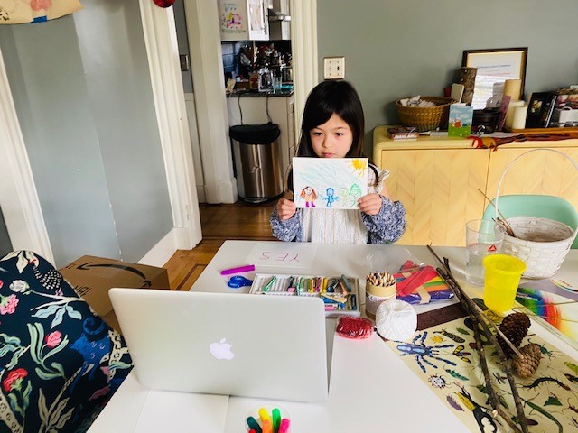 A young child holding up a crayon drawing to show on her laptop over a table full of colorful art supplies.
