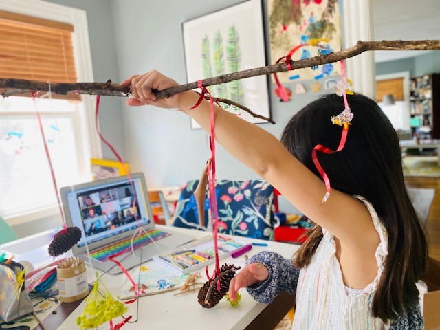 A young child raises a wood stick decorated with colorful streamers as she participates in a virtual workshop on her laptop.