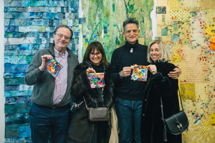A group of four adult visitors holding up their quilt square creations in front of a colorful, patterned tapestry background wall.