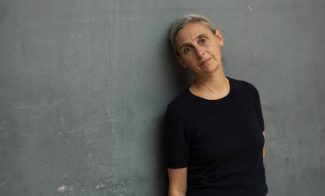 Anne Teresa De Keersmaeker, in a black top and blue jeans, leans against a gray wall.
