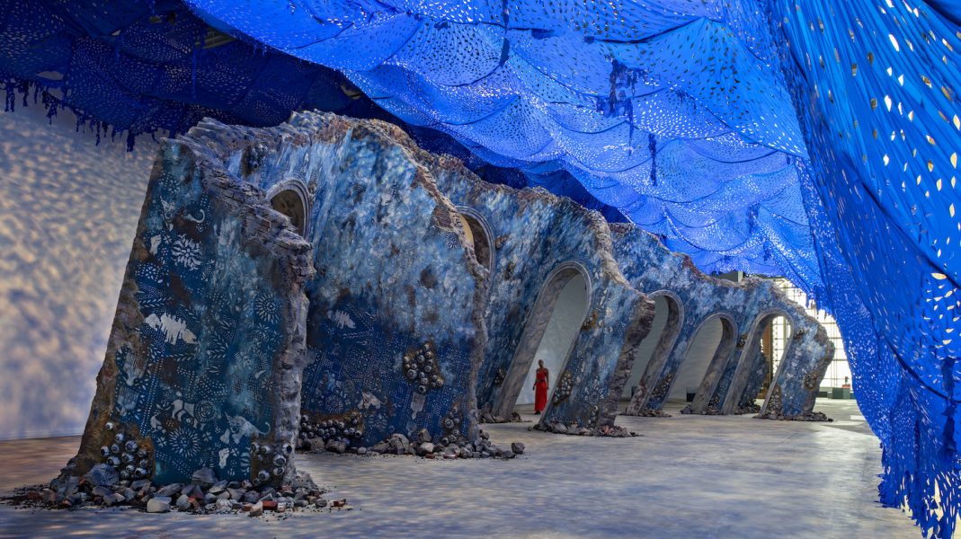 A woman in a red dress walks among a sculpture resembling an architectural ruin inside an industrial-style building. A bright blue perforated canopy creates a light-dappled effect on the floor and walls.