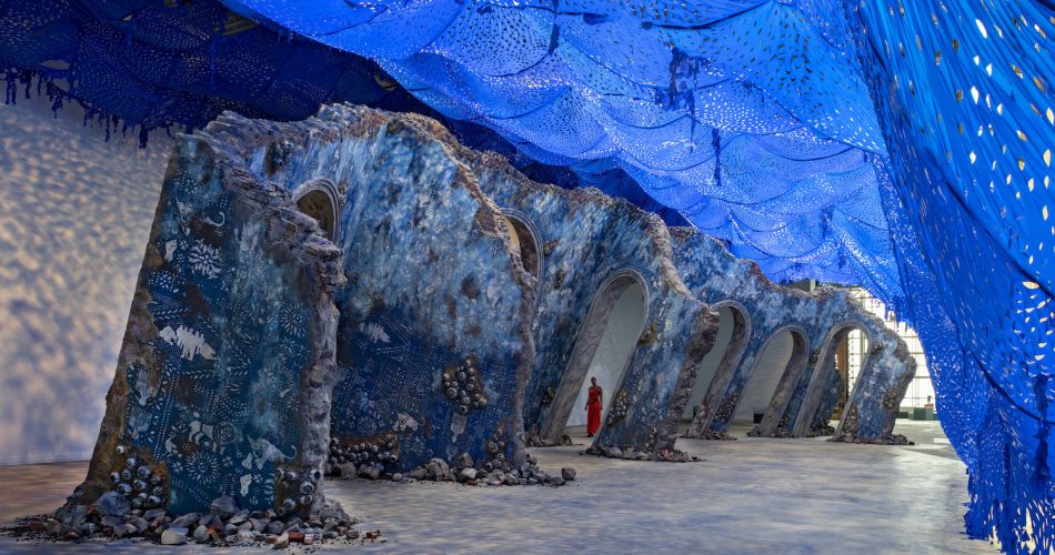 A woman in a red dress walks among a sculpture resembling an architectural ruin inside an industrial-style building. A bright blue perforated canopy creates a light-dappled effect on the floor and walls.