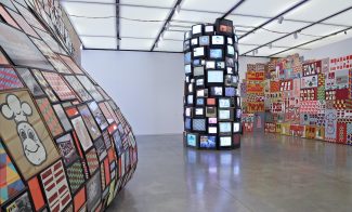 A monumental installation consisting of a towering sculpture made of television screens, a wall-sized mural, and a convex sculpture made of colorful framed graphics coming off the wall.