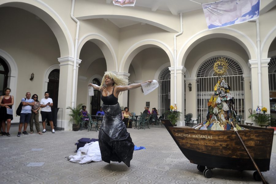 A medium-skinned woman wearing a blond wig performs in a courtyard. Her outstretched arms hold pieces of white paper with writing on them and she is jumping. A suitcase and blanket are behind her. About a dozen seated and standing spectators watch. 