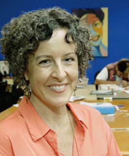 A headshot of Cynthia Campoy Brophy in a blue art room with teens drawing in the background.
