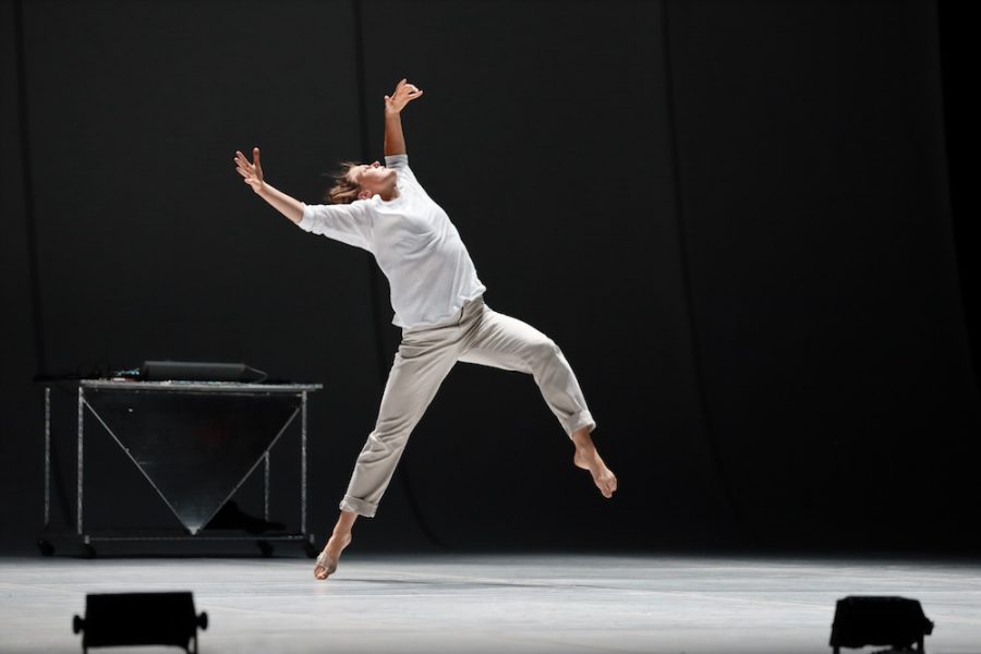 A man in a light shirt and pants seems to be caught mid-leap on a stage with a white floor and a black background 