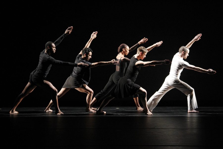 Four dancers in black and one in white lunge to the right with outstretched arms on a dark stage. 