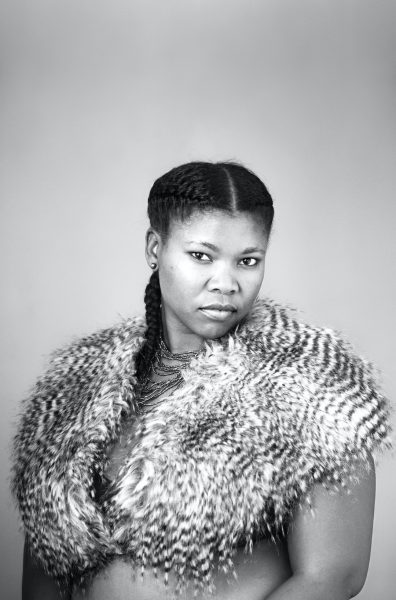 A black-and-white bust-length photograph of a person wearing a feathery capelet and beaded necklaces