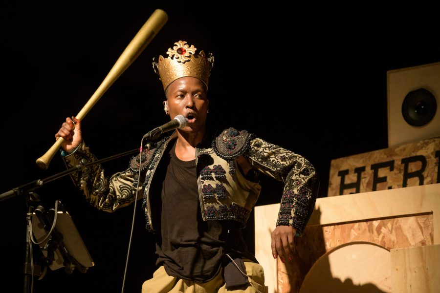 A dark-skinned woman wearing a crown holds up a baseball bat in one hand. She's on a stage set and speaking or singing into a microphone on a stand. 