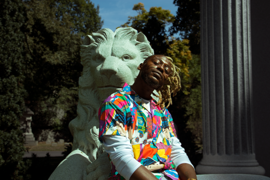 A photo portrait of Cliff Notez, an African-American musician in a colorful shirt leaning against a lion statue with a roman column and greenery in the background.