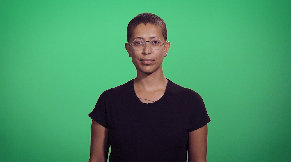 A video still of a medium-skinned person with glasses and short hair, shown from the torso up, facing the viewer against a green background.