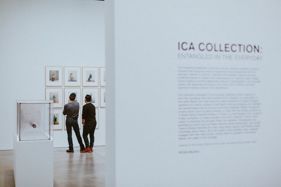 A view through a doorway shows two men looking at a wall of framed photographs from behind. In the foreground is a wall with text that says ICA Collection.