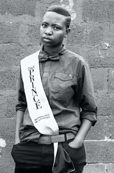 A black-and-white photograph of a person standing against a wall wearing a white sash with the words 