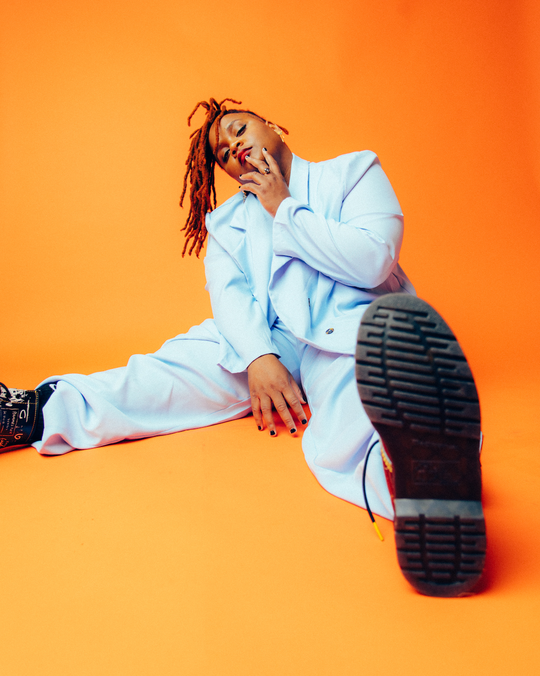 An African-American artist with red dreads wearing a pale blue suit and boots in a sit down pose in front of an orange backdrop.