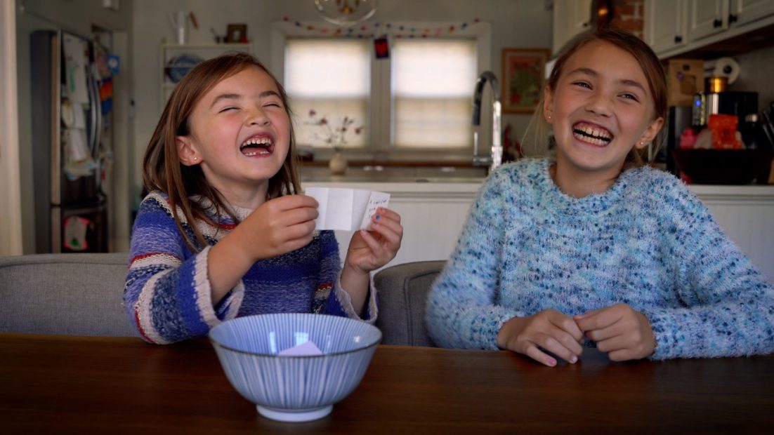 Two children smiling and sitting at the dining table in their home, with one holding up a piece of paper drawn from a ceramic bowl.