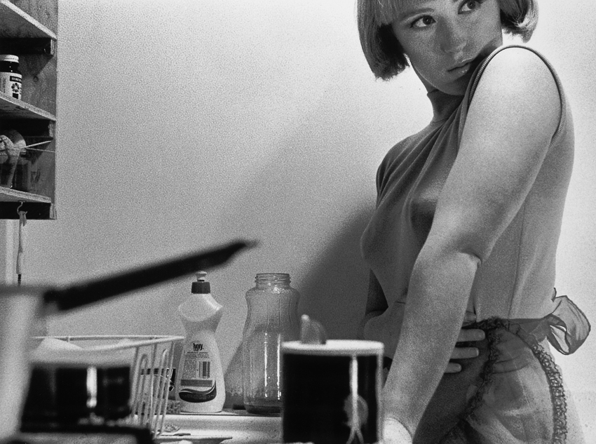 A black-and-white photograph of the artist posing in a small, cramped kitchen, looking over her shoulder. She wears a sleeveless top and dramatic eye makeup and the image is cropped to include dishes and dish detergent but cut off the top of her head.