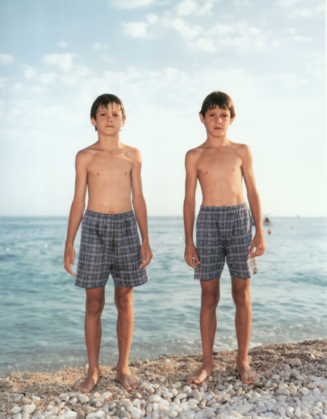 A color photograph of two light-skinned young boys in plaid swimsuits standing side-by-side at the edge of the water on a rocky beach.