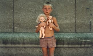A color photograph of a tanned and dirty young boy with blond hair wearing only shorts and  clutching two unclothed dolls against his bare chest.