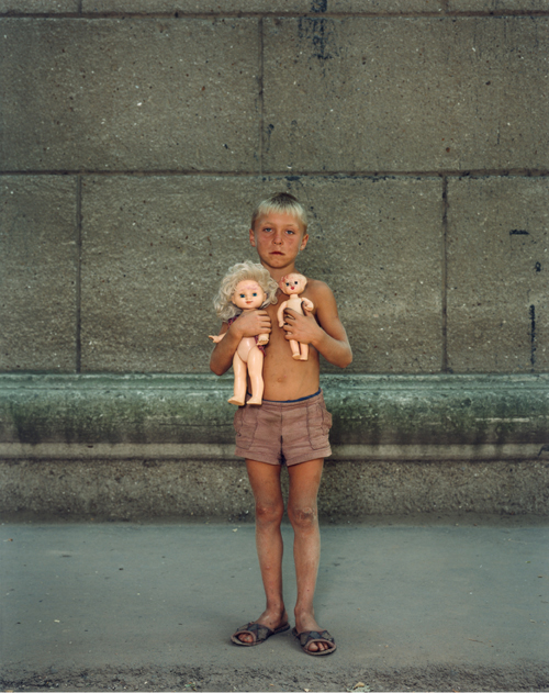 A color photograph of a tanned and dirty young boy with blond hair wearing only shorts and  clutching two unclothed dolls against his bare chest. 