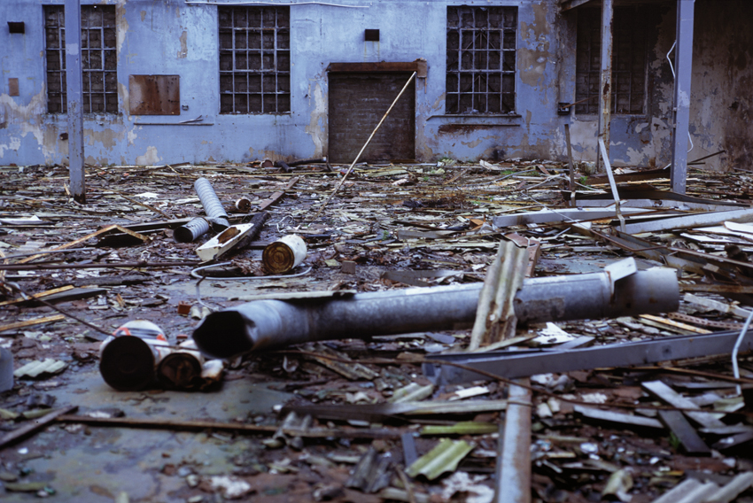 A color photograph of an industrial yard covered in debris with a dilapidated building in the background. 
