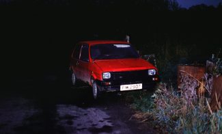 A color photograph of an abandoned, red car on the side of a dark road at night.