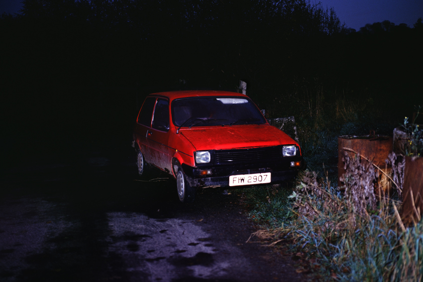 A color photograph of an abandoned, red car on the side of a dark road at night.