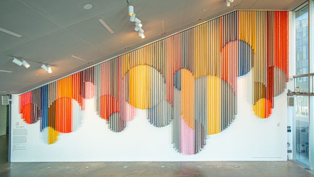 A monumental hanging wall sculpture made from bands of colorful coated mesh fabric which drape in various lengths to create series of interlocking circular forms, and installed in an empty spacious lobby. 