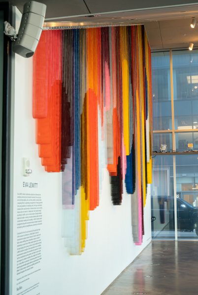 A side-angle view of a monumental hanging sculpture on a wall that's made up of bands of colorful coated mesh fabric which drape to create a series of interlocking circular forms, in an empty lobby space.